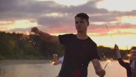 The-young-student-in-black-t-shirt-is-dancing-with-big-bengal-lights-on-the-sand-beach-with-his-friends.-This-is-crazy-summer-evening-on-the-open-air-party.